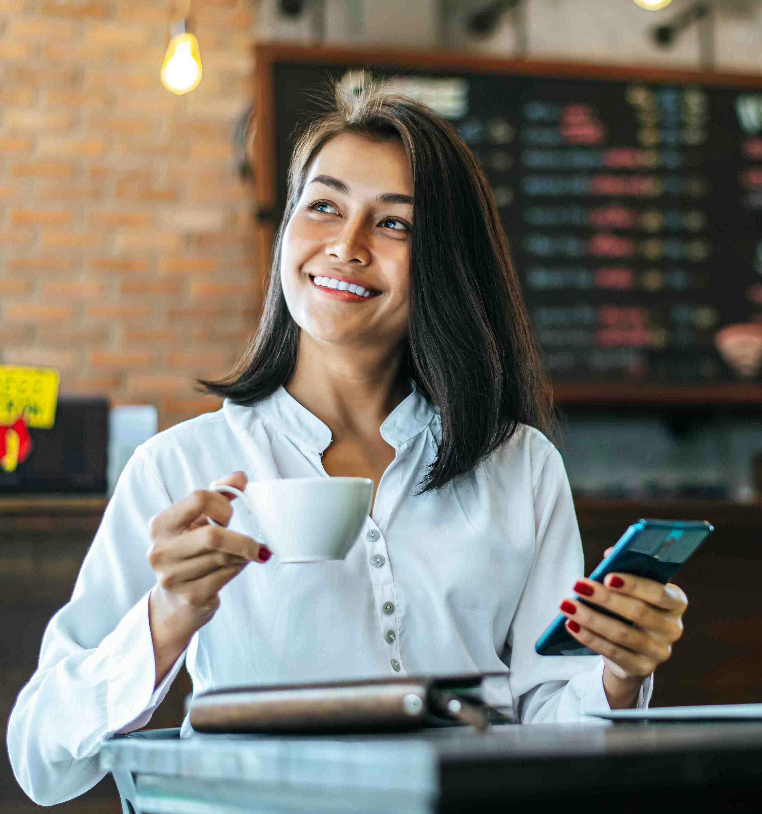 Mulher representando a dona de um estabelecimento segurando um celular enquanto toma um café